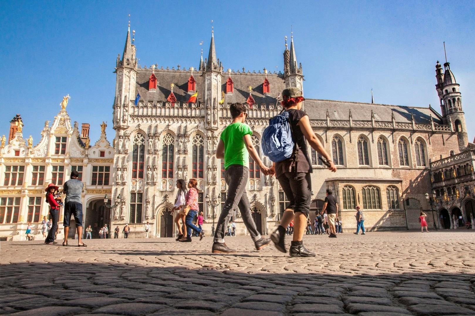 People walk and take photos in a large square with historic Gothic buildings under a clear blue sky.