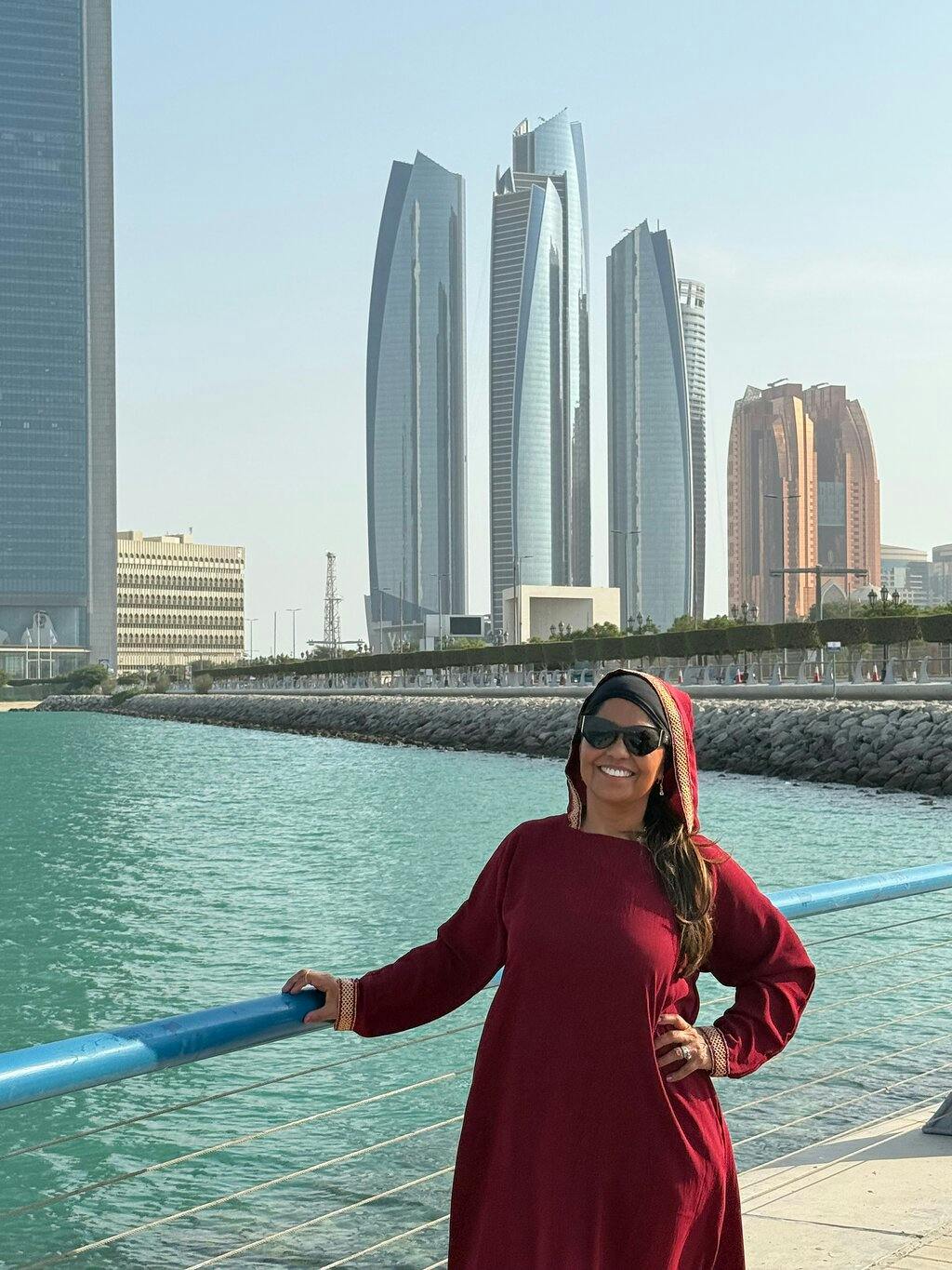 A woman in a red outfit and sunglasses smiles by a waterfront with modern skyscrapers in the background.