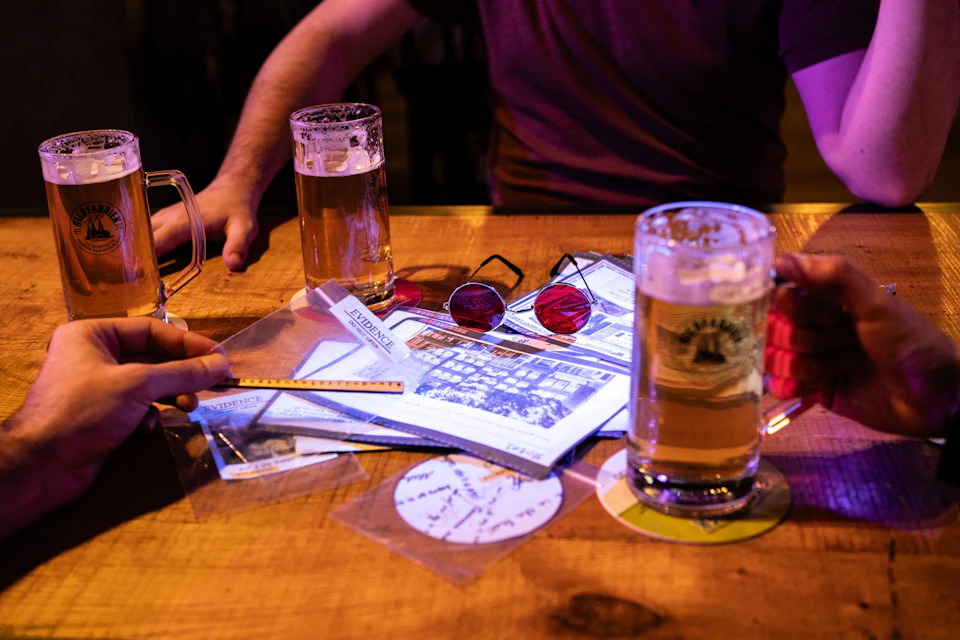 Two people sitting at a table with beer, papers, red-tinted glasses, and a ruler.