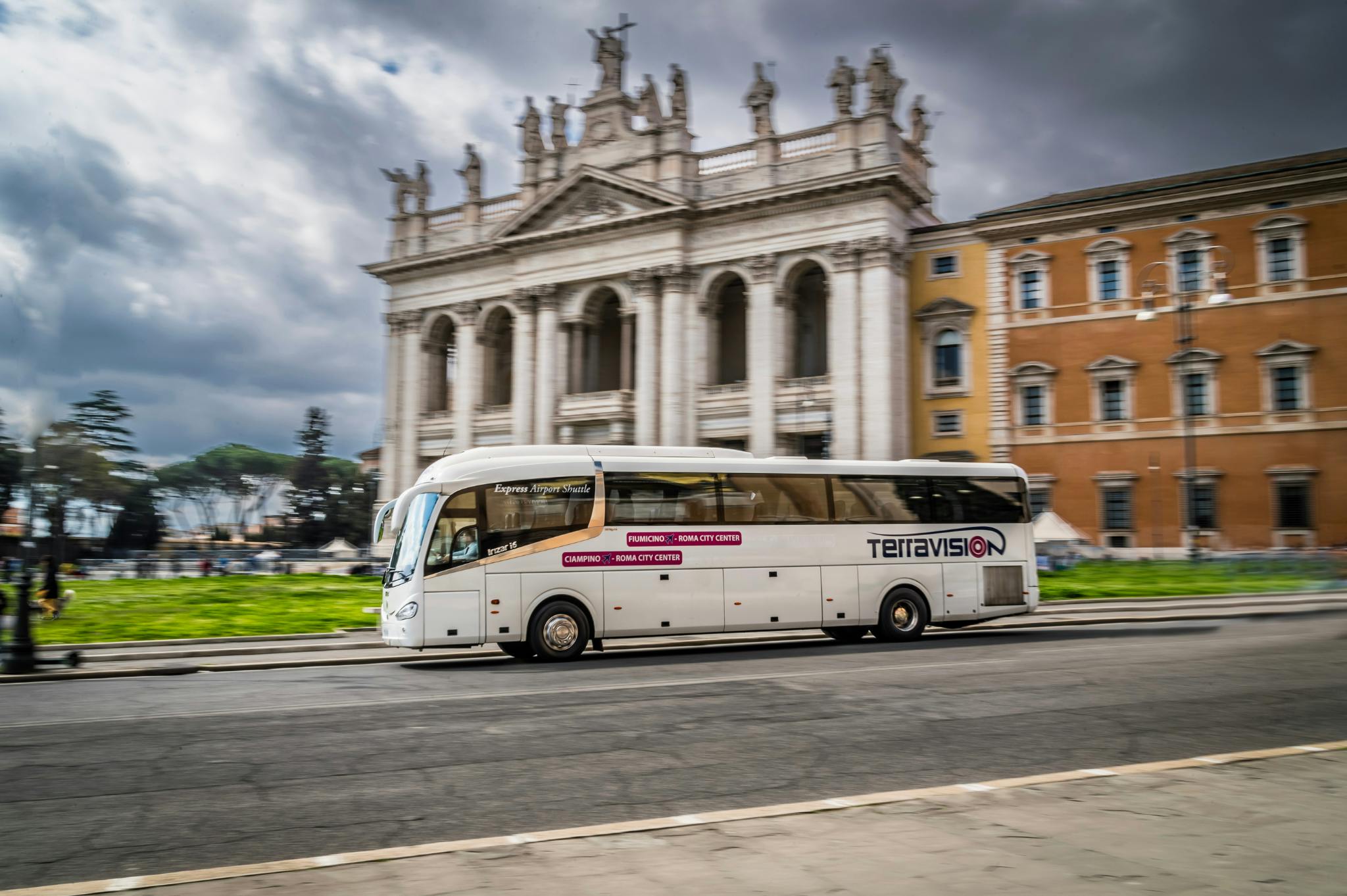 A Teravision coach bus travels past a historic European building with columns and statues. Cloudy sky in the background.