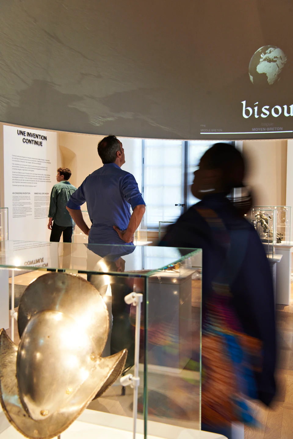 People examining exhibits in a museum room with "Une Invention Continue" text on the wall and glass display cases.