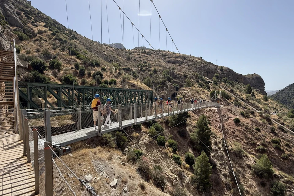People walking on a suspension bridge over a canyon, surrounded by dry hills and vegetation under a sunny sky.