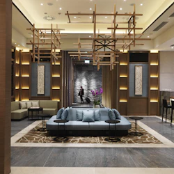 En modern hotellobby med en central sittgrupp, dekorativa träpaneler, hängande armaturer och en enda person som går.