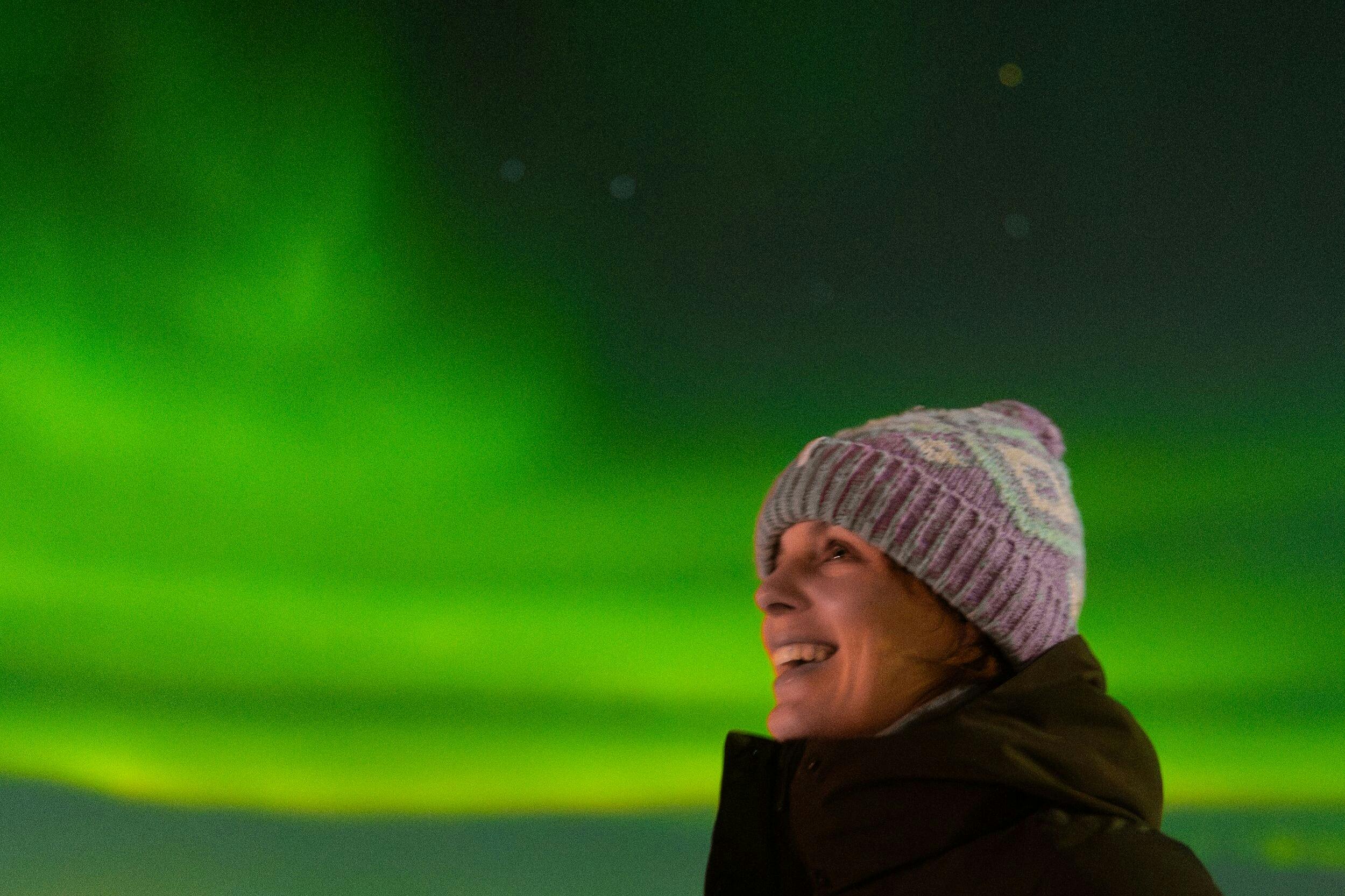 A person in a winter hat and coat smiles with green northern lights visible in the night sky in the background.
