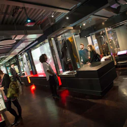 Människor utforskar utställningar i ett modernt museum med glasfodral och interaktiva skärmar under industriell belysning.