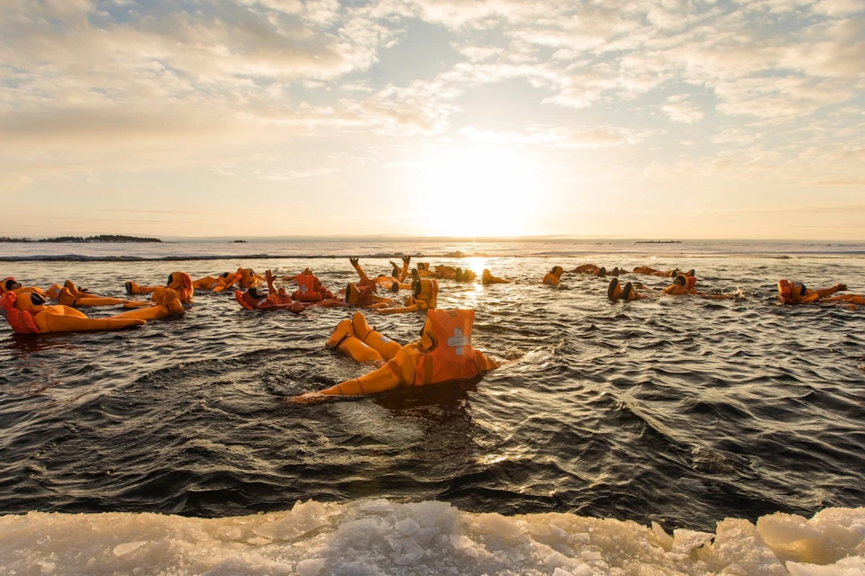 People wearing orange survival suits floating on a cold water surface under a partly cloudy sky with sunlight.