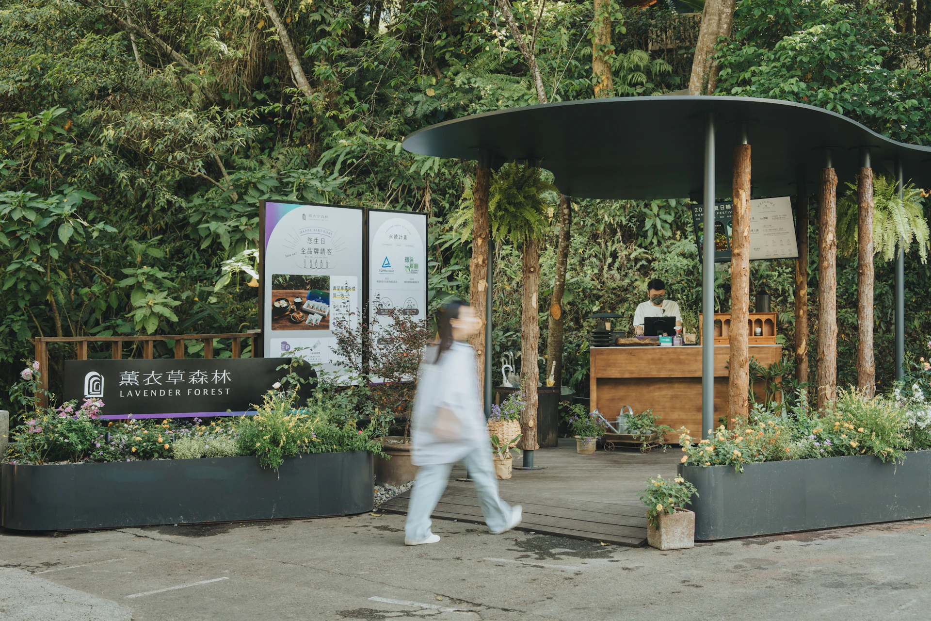 An outdoor reception area in a forest with a person walking past, a staff member working, and informational signage visible.