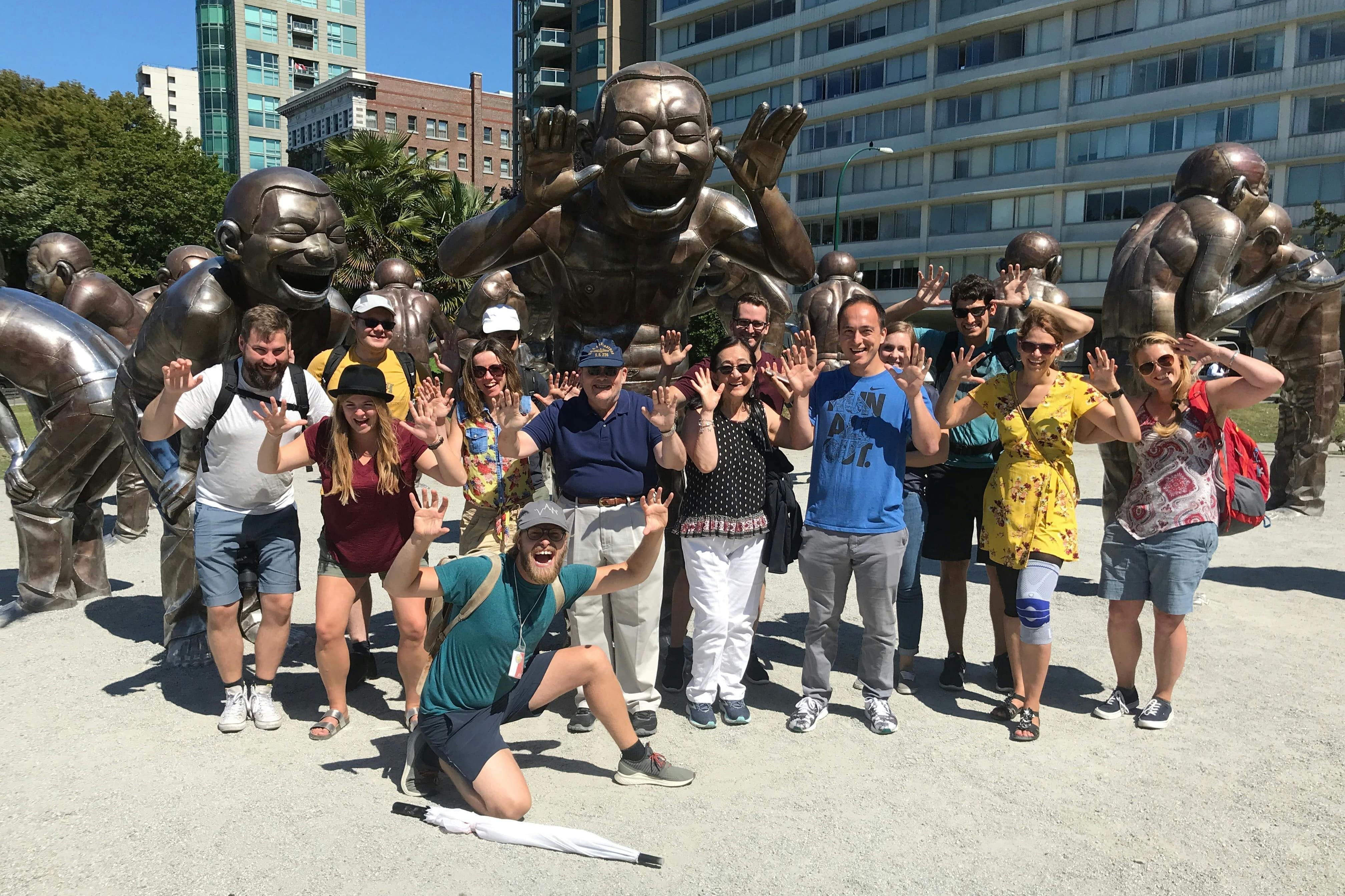 Skupina lidí pózujících před bronzovými smějícími se sochami ve venkovním městském prostředí za slunečného dne.