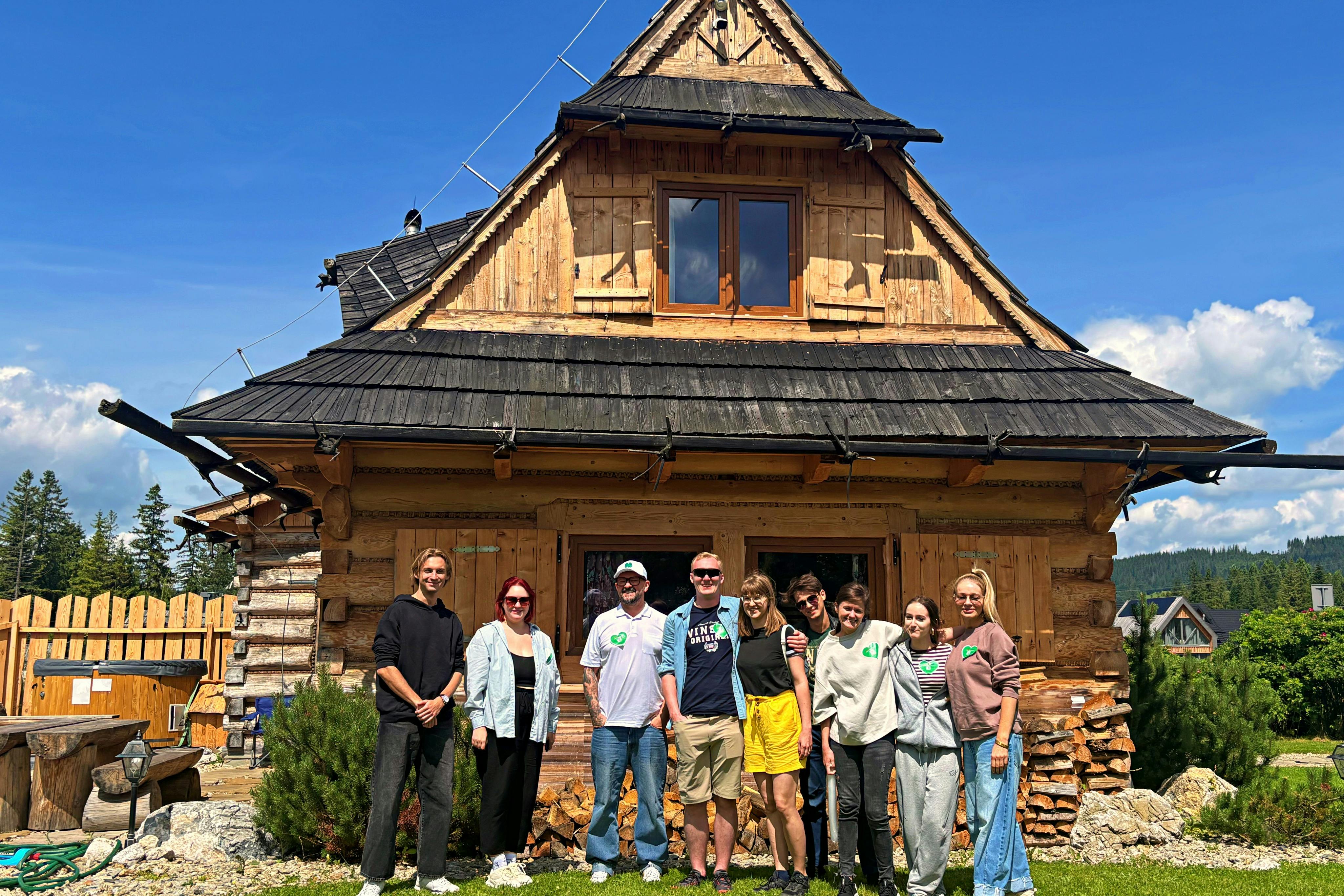 화창한 날씨에 푸른 잔디와 흐린 하늘을 배경으로 7명의 일행이 소박한 목조 주택 앞에 서 있습니다.