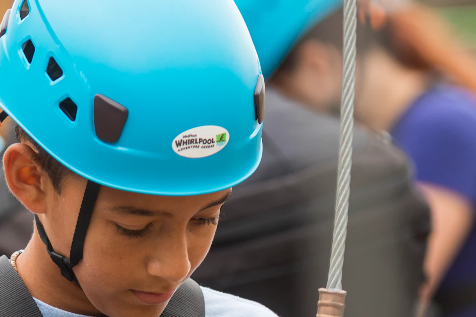 People wearing blue helmets and safety harnesses are attaching clips to a wooden post while preparing for a zipline or ropes course.