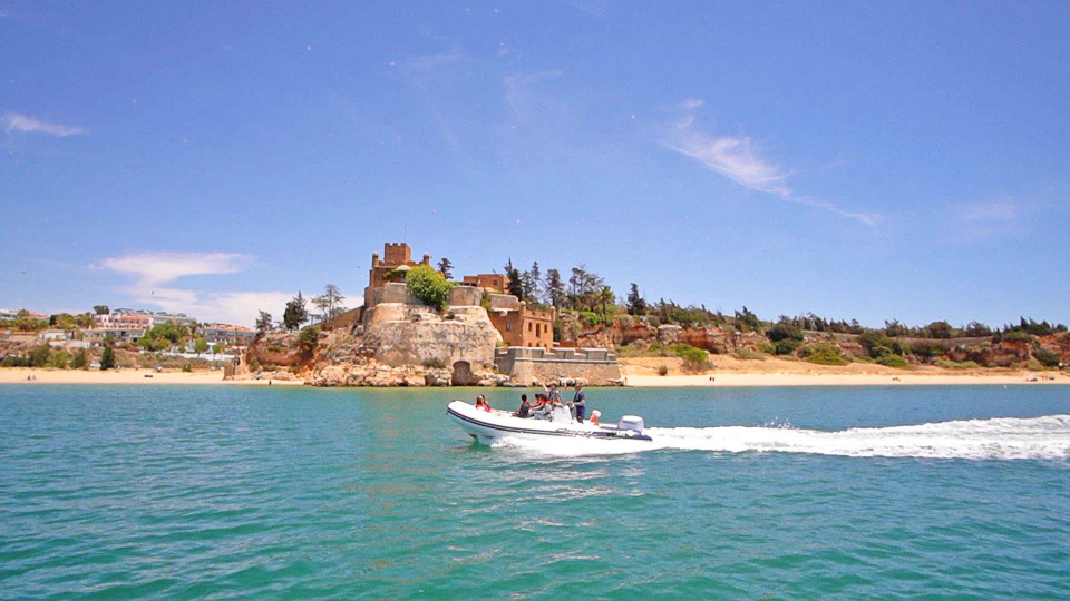 A small motorboat with passengers travels on blue water near an old fortress on a sunny day.