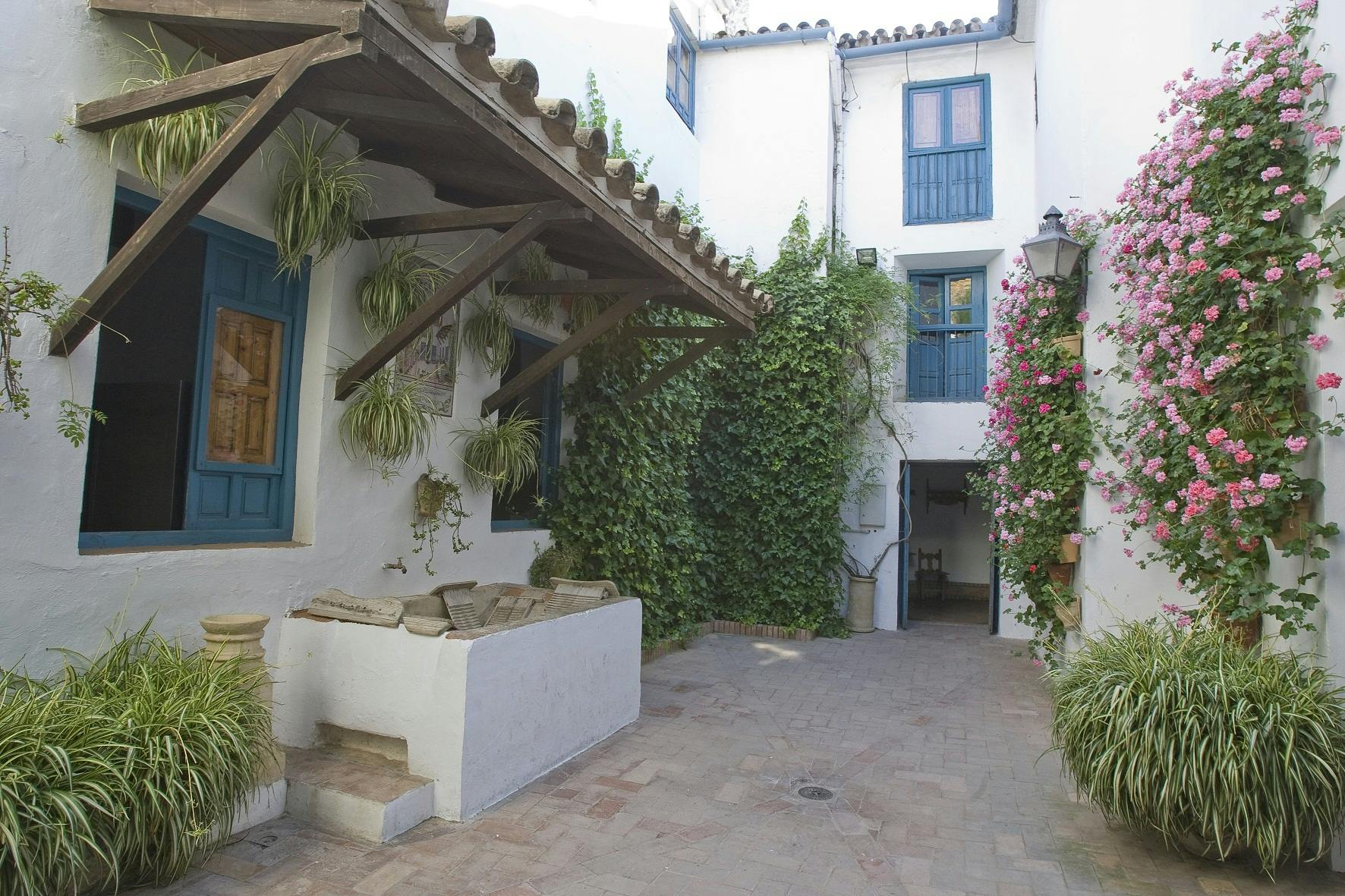 Courtyard with white walls, blue windows, wooden awning, hanging plants, and ivy-covered wall. Pink flowers and lantern on the right.