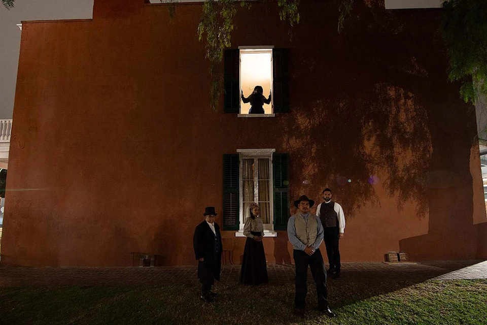Four people in period clothing stand in front of a building at night; a figure is silhouetted in a lit upper window.