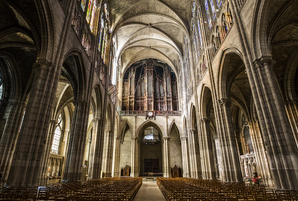 Interior of a large cathedral featuring rows of wooden pews, vaulted ceilings, stained glass windows, and a grand organ.