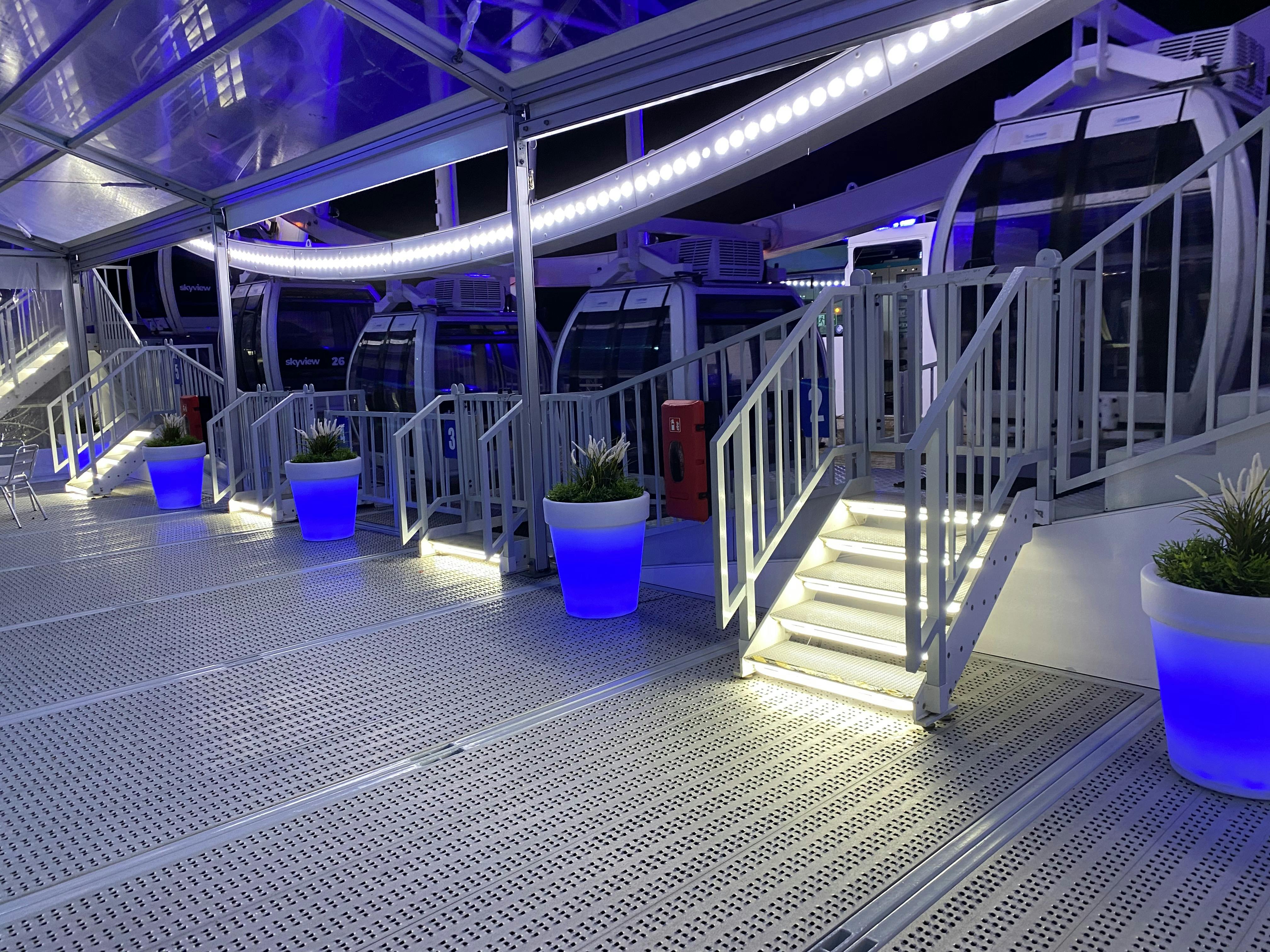 通往观景轮玻璃封闭吊舱的照明楼梯。 人行道上种有蓝色灯光的盆栽植物。