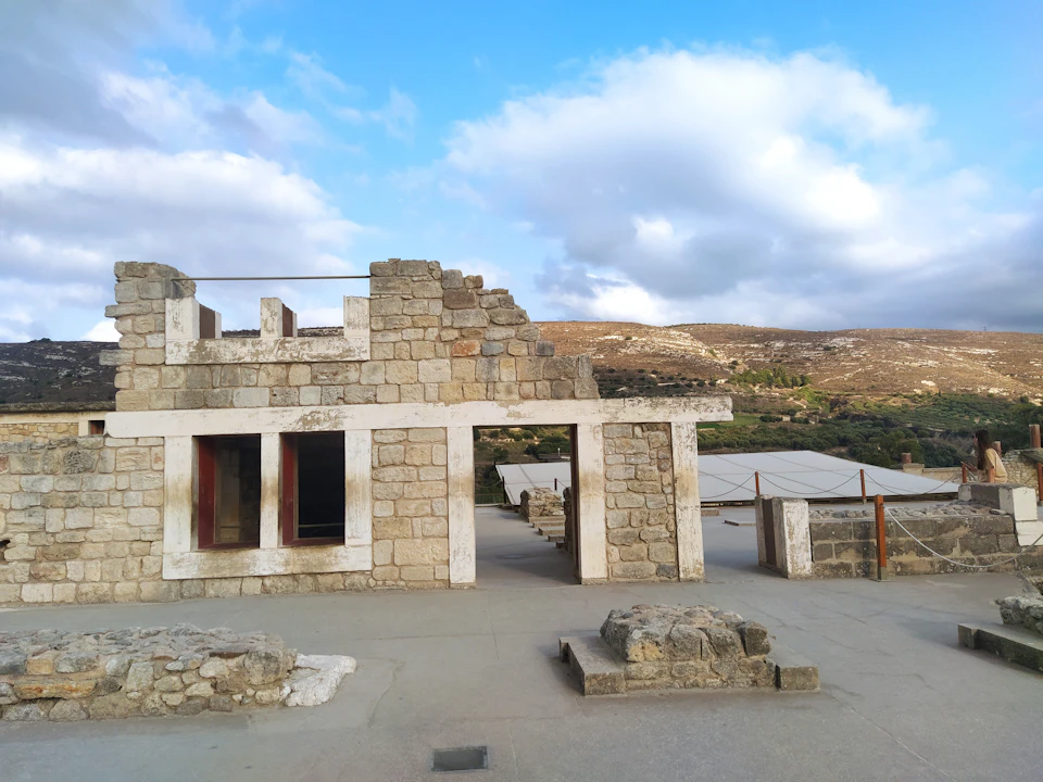 Ancient stone building ruins under a partly cloudy sky, with hills in the background and some modern structures nearby.