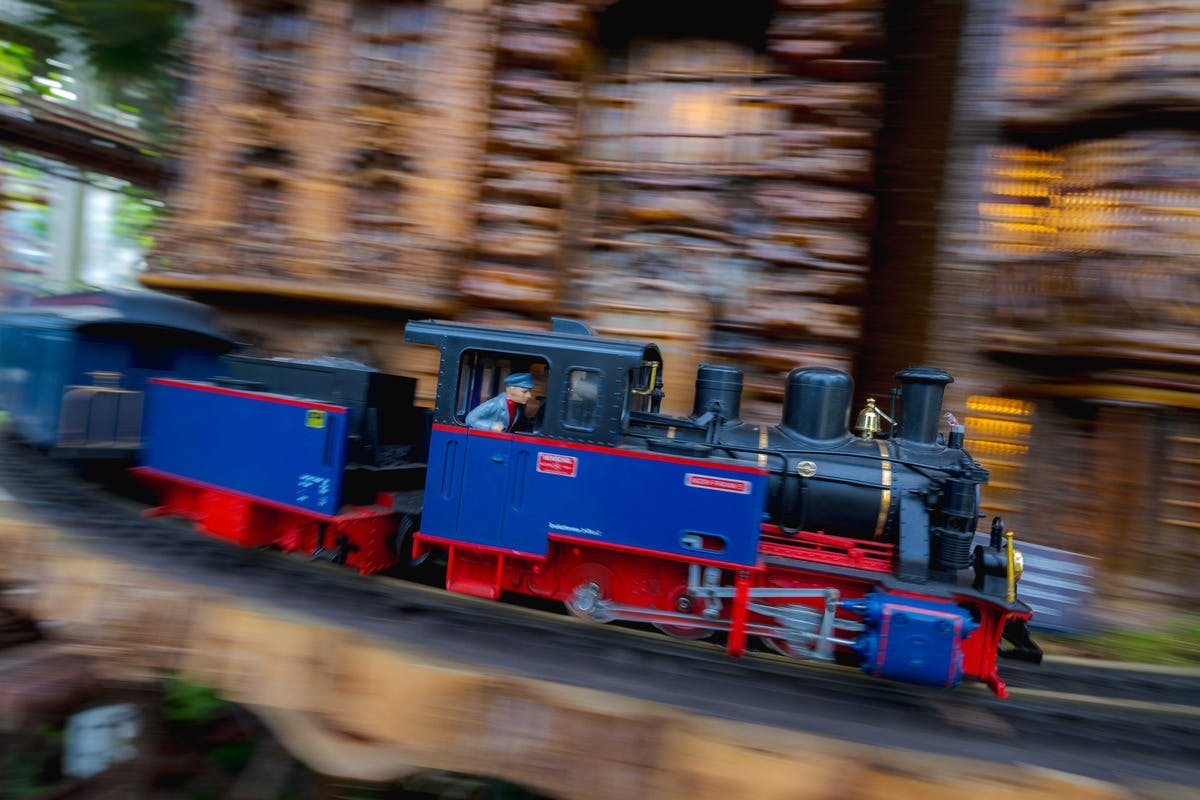 Et miniatureblåt og rødt damptog i bevægelse med en sløret gyldenbrun bygning i baggrunden.