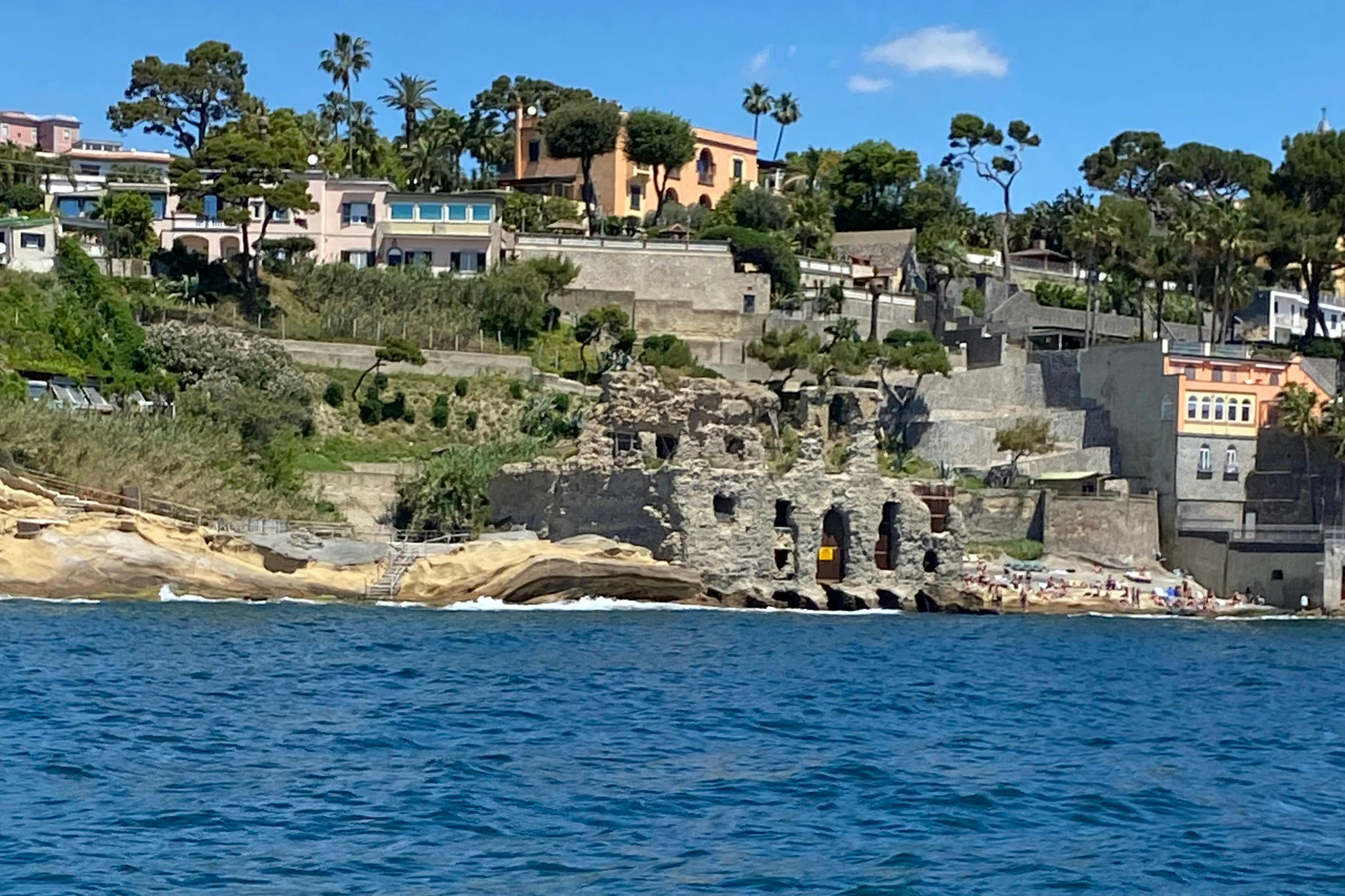 Vista costera de casas en la ladera, ruinas cerca de la orilla y bañistas dispersos, con el mar azul en primer plano y el cielo despejado por encima.
