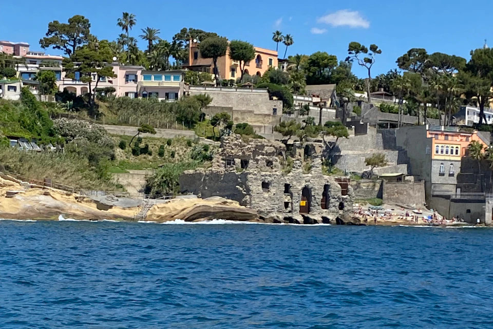 Coastal view of hillside homes, ruins near the shore, and scattered beachgoers, with blue sea in the foreground and clear sky above.
