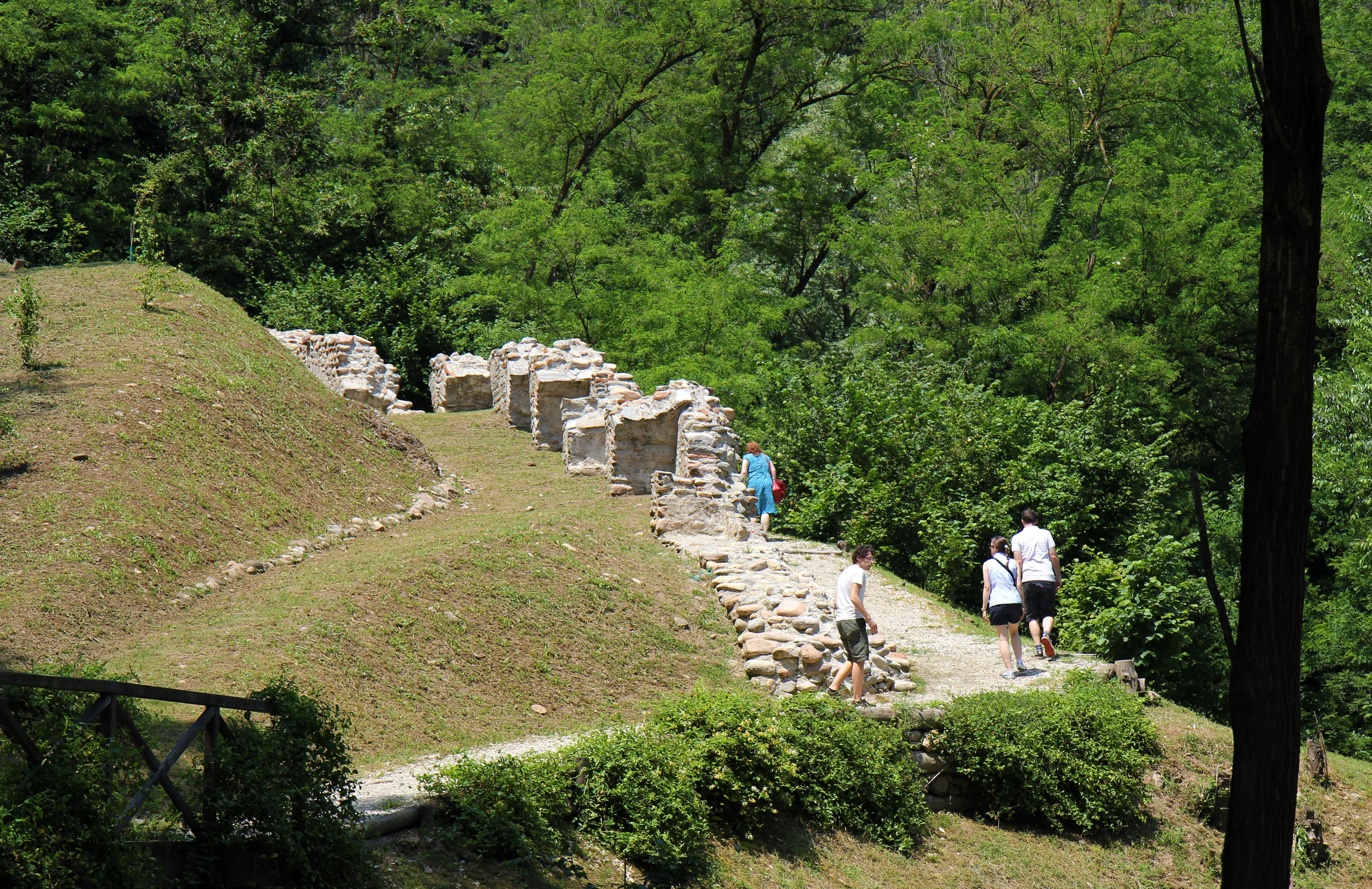 People walking along a path near ancient stone ruins in a lush, green forest.