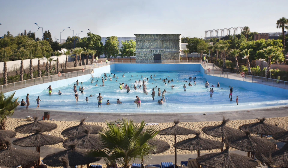 People enjoying a wave pool, surrounded by palm trees and shaded areas with straw umbrellas and lounge chairs.
