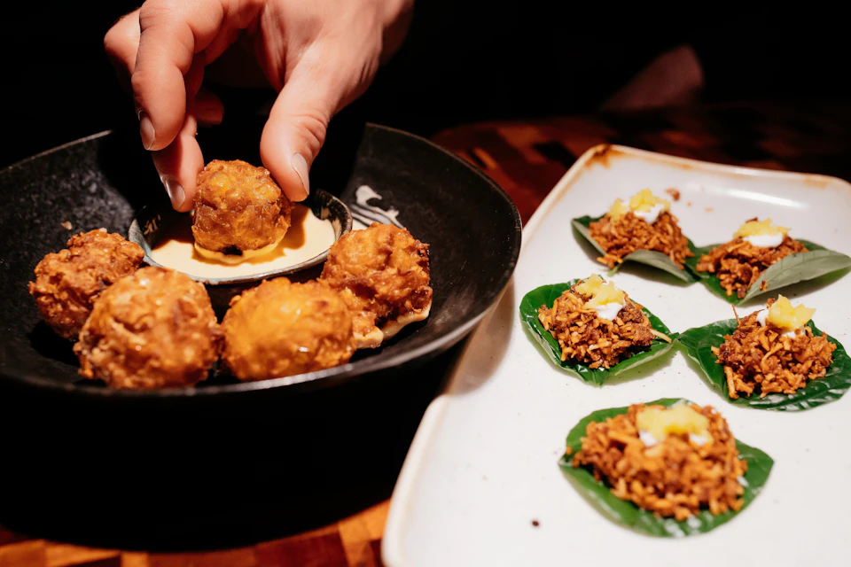 A hand placing a fried appetizer in a black dish, next to a plate of leaf-based snacks topped with crispy toppings.