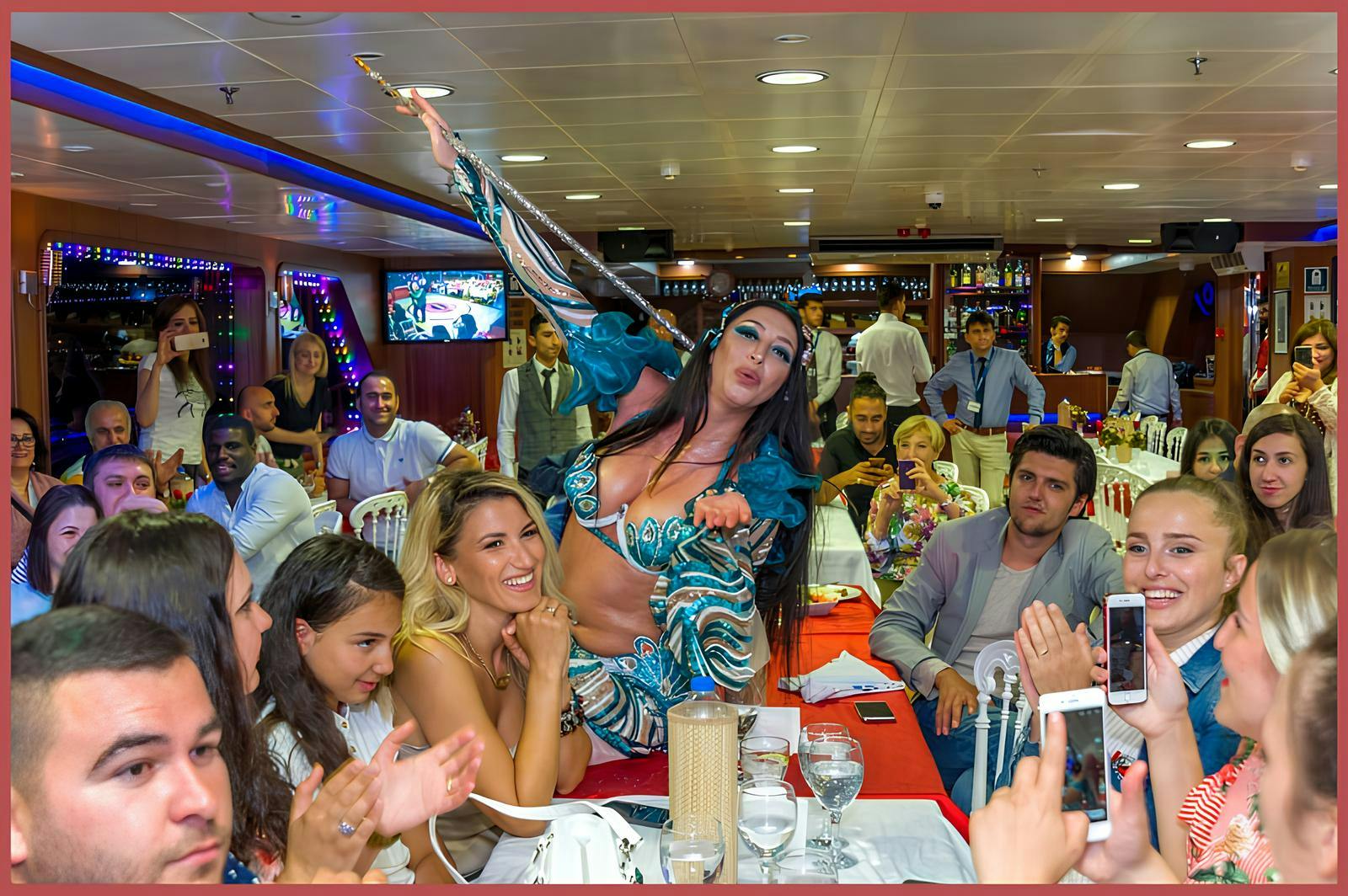 一位穿着充满活力的服装表演者在拥挤的节日室内环境中在桌子上跳舞。 周围的人在拍手和拍照。