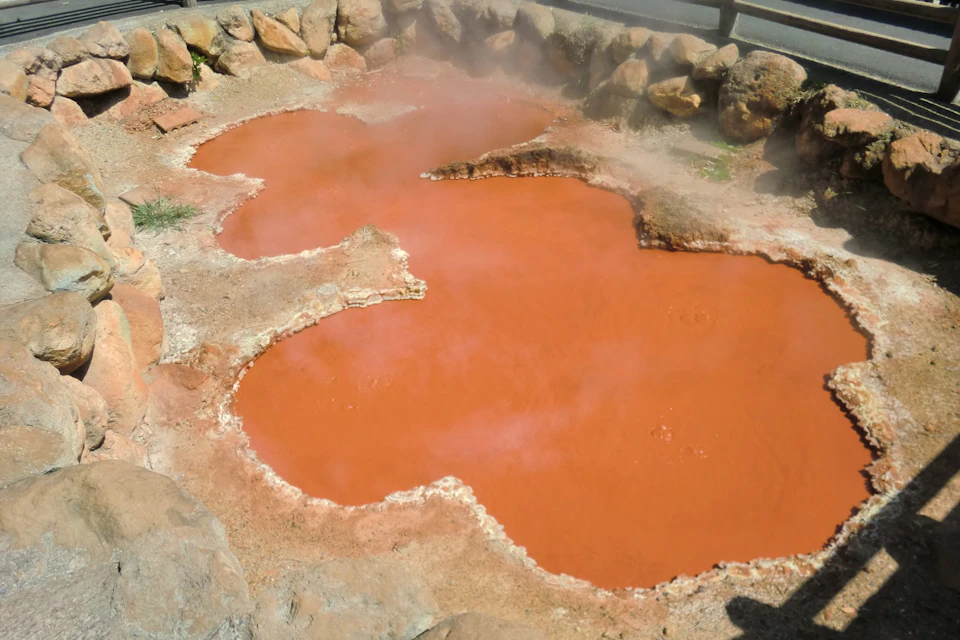 A natural hot spring with orange-red water, surrounded by rocks and emitting steam, under an outdoor setting.
