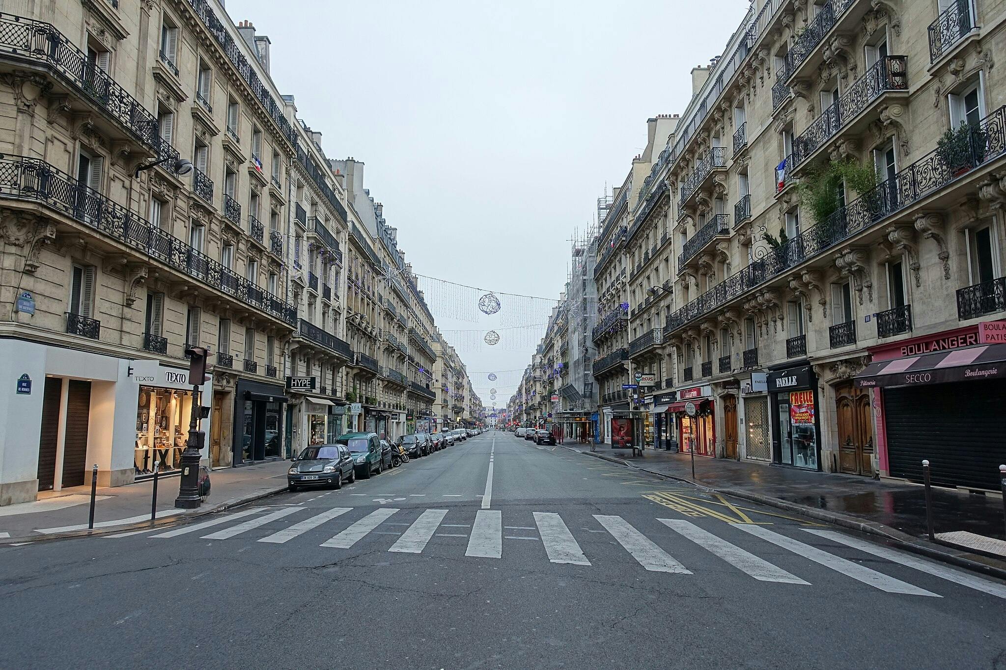 Urban street scene with classic European architecture, lined with shops and parked cars. Decorative lights hang above the road.