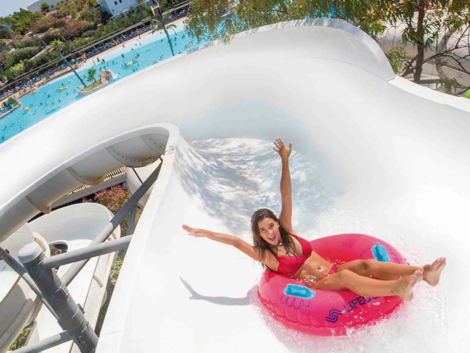 A woman in a red bikini is sliding down a water slide on a pink inflatable ring, smiling with arms raised. A pool is visible in the background.