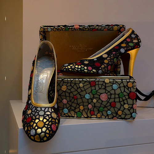 Par höga klackar med guldklackar och ett färgglatt mosaikmönster, som visas med en matchande handväska på vita hyllor.