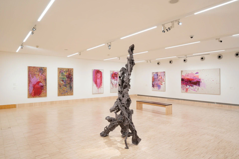 Art gallery with abstract paintings on the wall, a large sculptural piece in the center, and a wooden bench near the back wall.