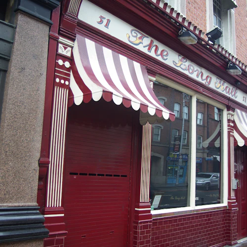 En skyltfönster med röd- och vitrandiga markiser, en röd fasad, stort fönster och namnet ”The Long Hall” som visas ovan.