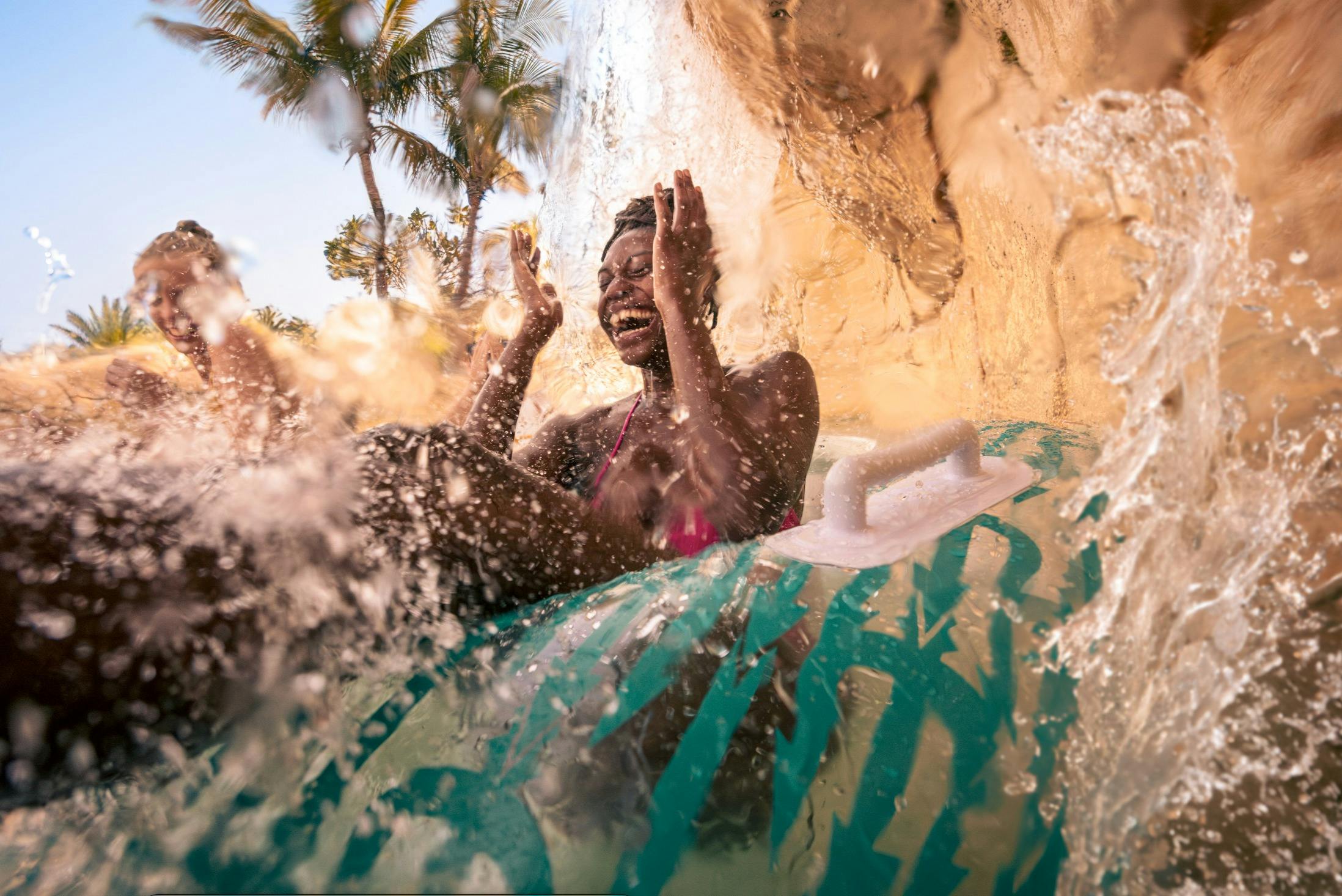 Человек на надувной трубе, наслаждающийся водной горкой под водопадом, с пальмами на заднем плане.