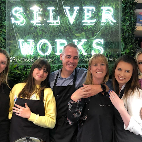 Six people wearing black aprons pose in front of a "SILVER WORKS" neon sign with greenery, smiling and showing rings.