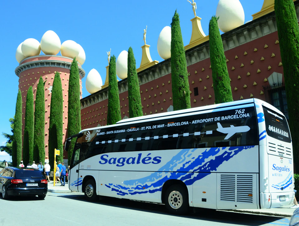 Tour bus parked beside a unique building with egg-shaped sculptures and cypress trees. People are near the bus.