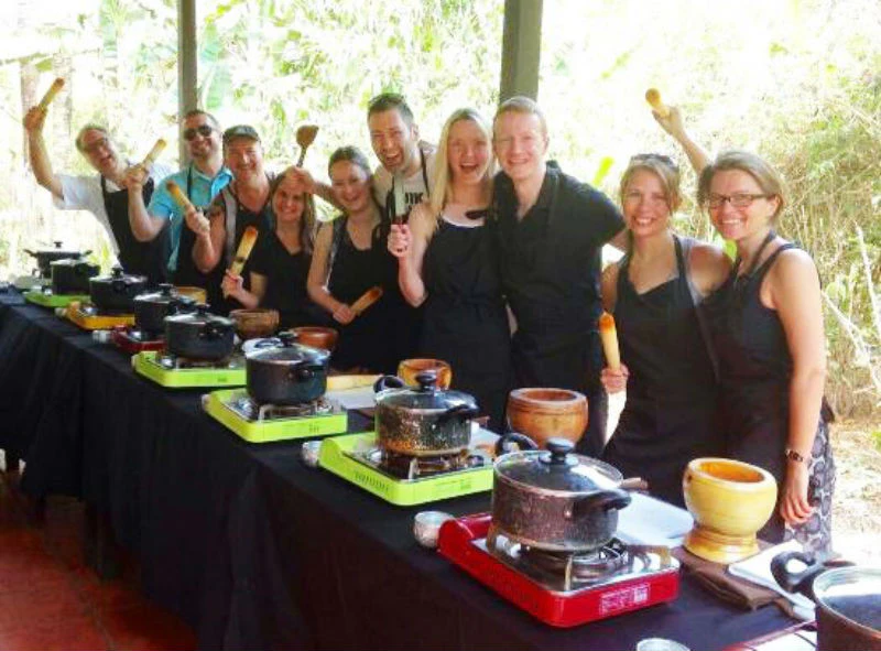 A group of people in black aprons smile while standing at a table with cooking pots and utensils in an outdoor setting.