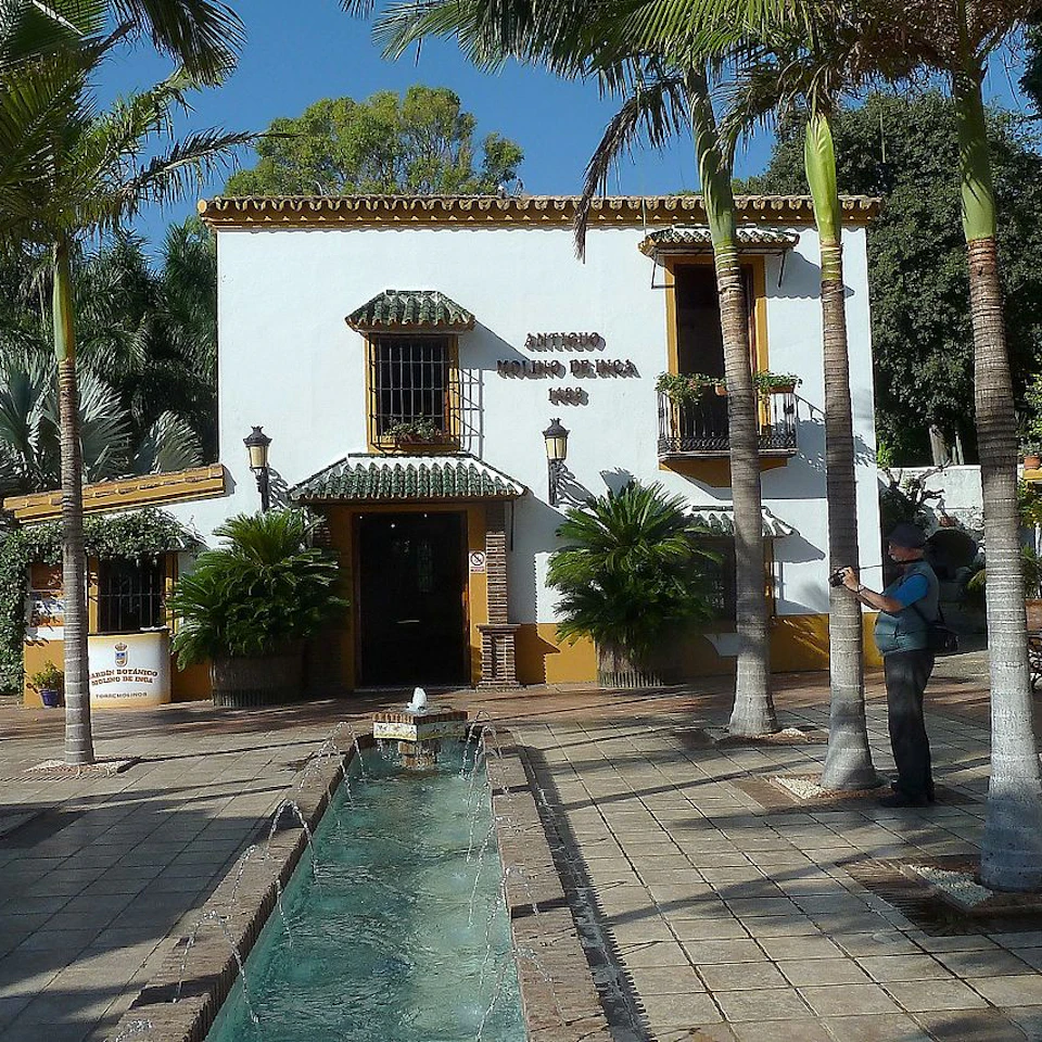 A white two-story building with green trim and a tiled courtyard featuring a narrow water channel, surrounded by palm trees.