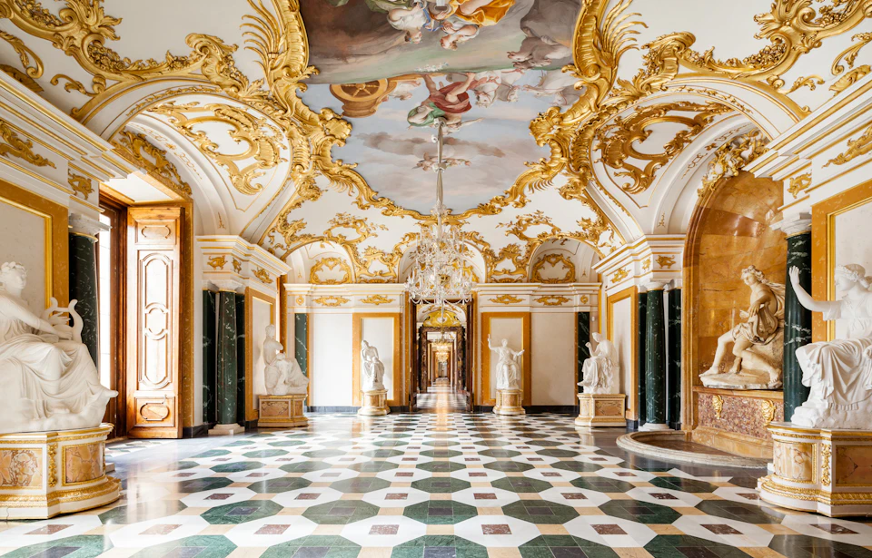 Ornate palace hallway with intricate ceiling frescoes, gold detailing, marble statues, and a chandelier. Geometric tiled floor.
