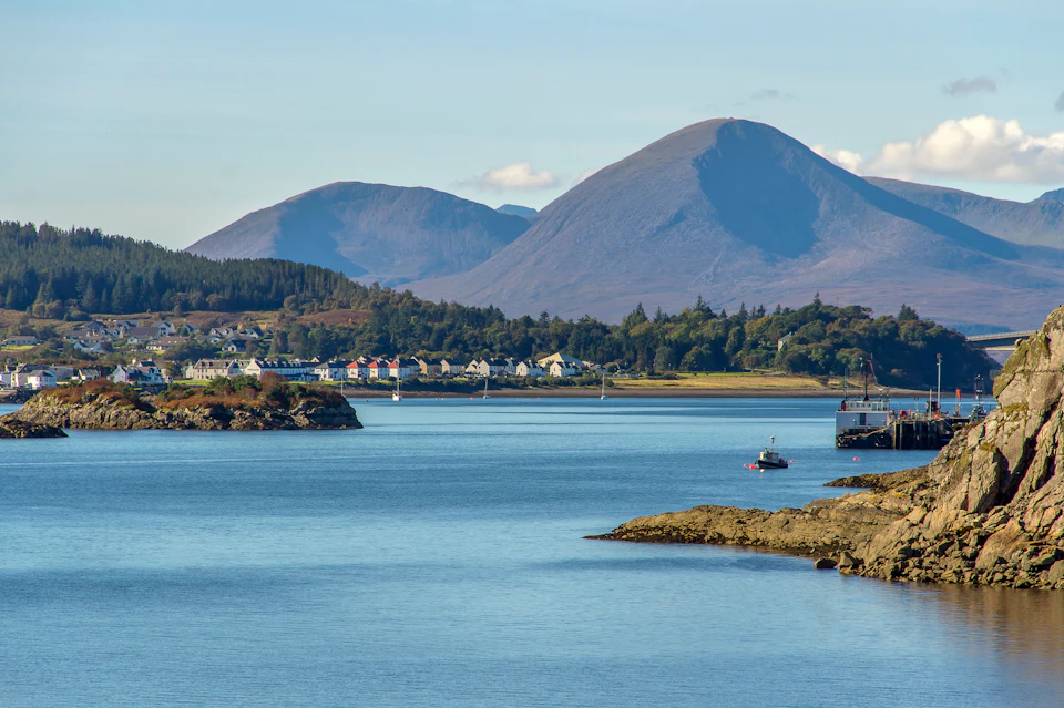 Coastal view with calm water, houses nestled among trees in the distance, and large mountains under a clear blue sky.