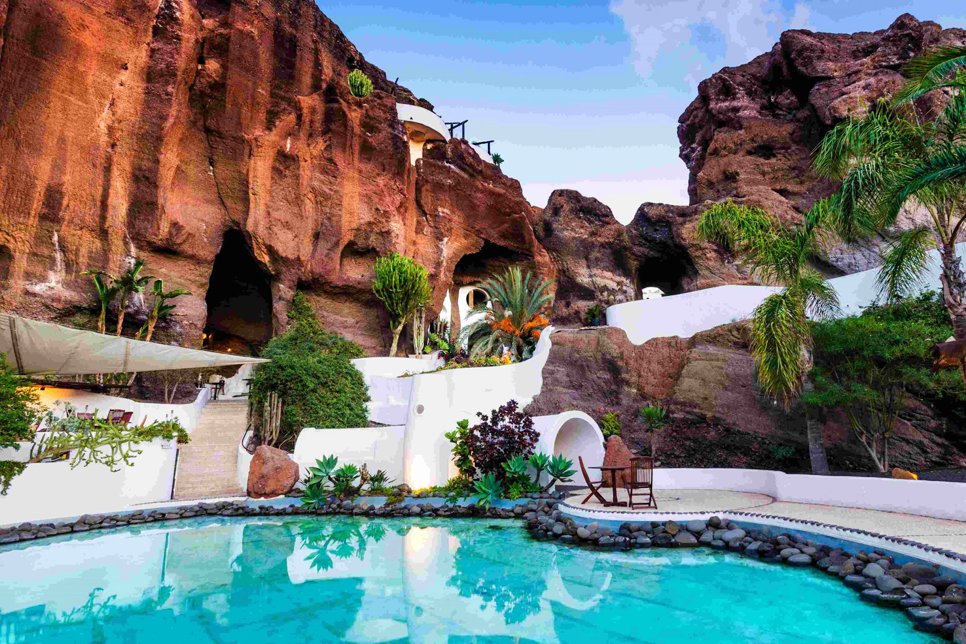 A serene pool with reflections of lush plants and white buildings carved into a rocky hillside under a partly cloudy sky.
