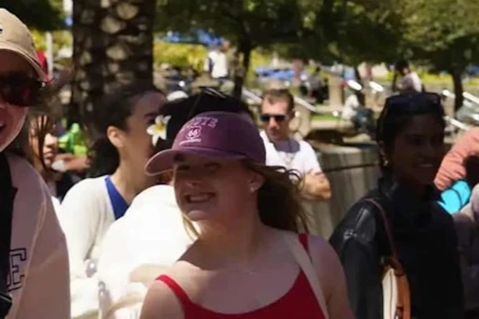 Three people stand outdoors, smiling at the camera. They are in casual clothing, enjoying a sunny day with palm trees and buildings behind them.