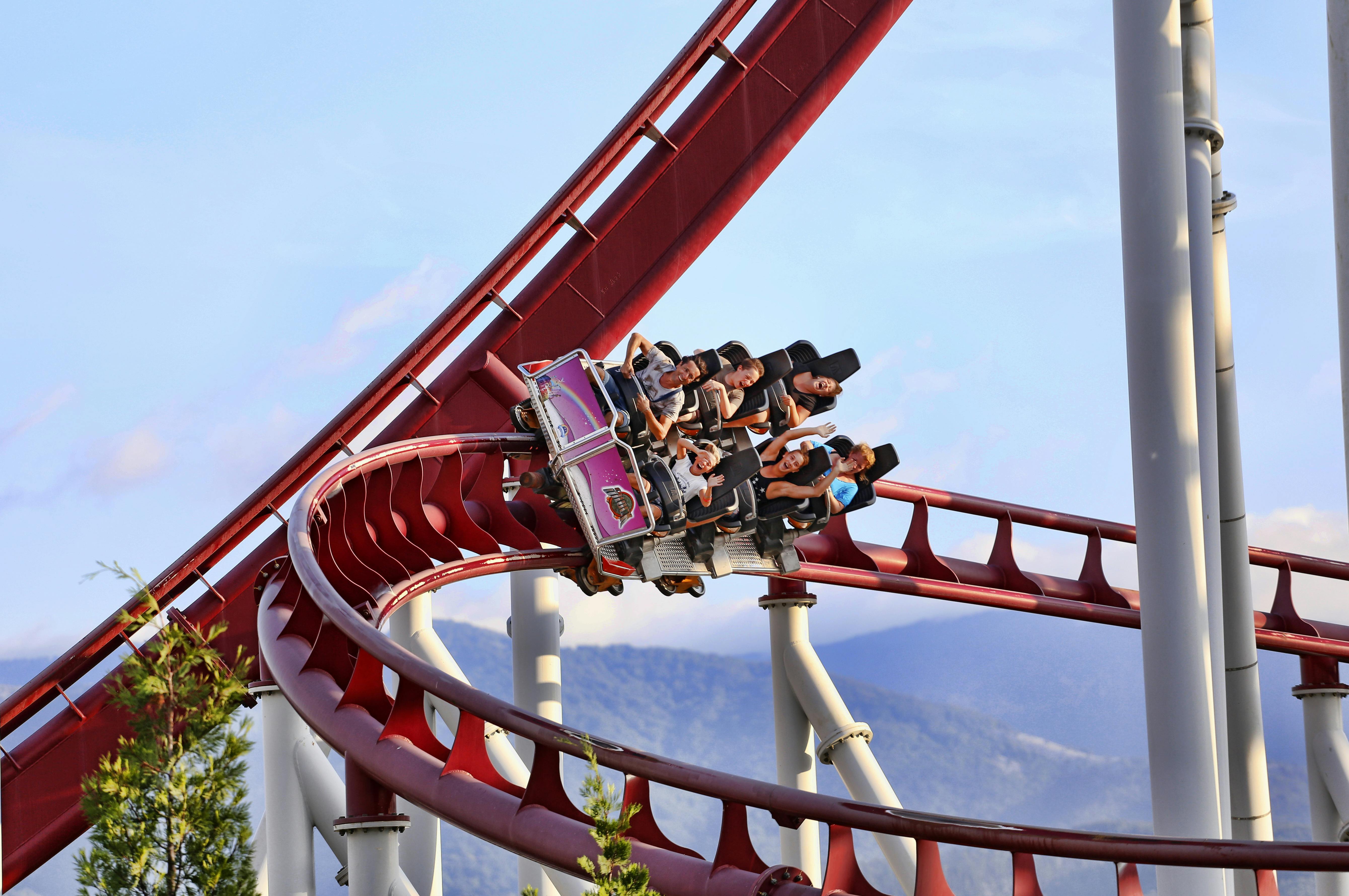 人们乘坐带有红色轨道的过山车，向一侧倾斜。 背景显示晴朗的天空和遥远的山丘。