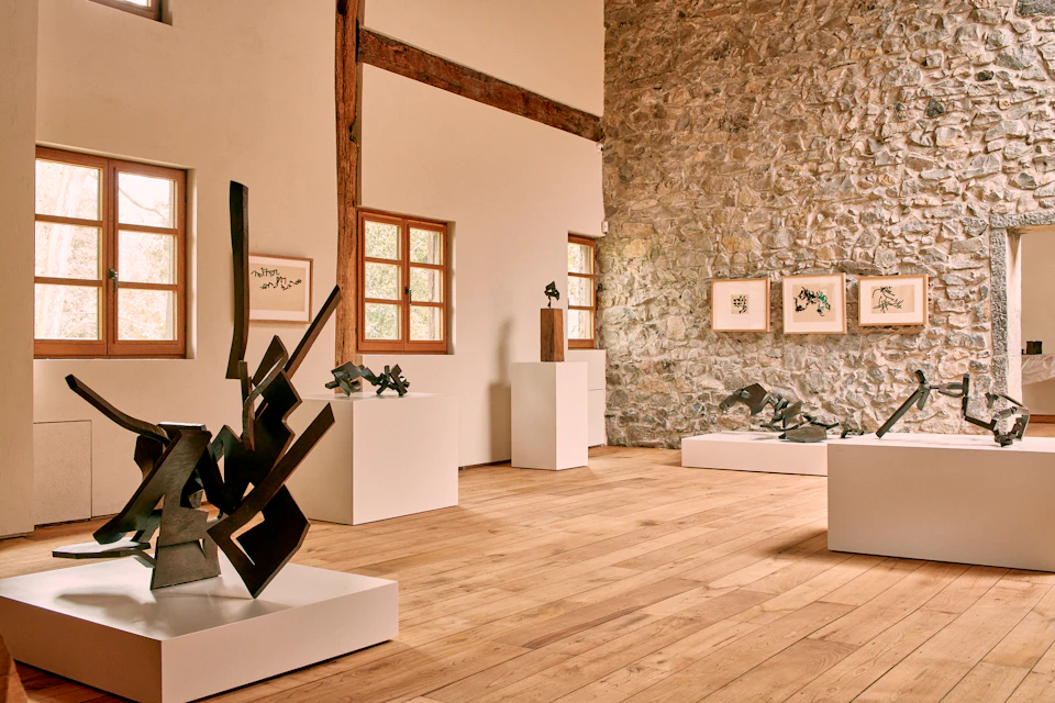 Art gallery interior with modern sculptures on white pedestals, framed artworks on a stone wall, and wooden flooring.