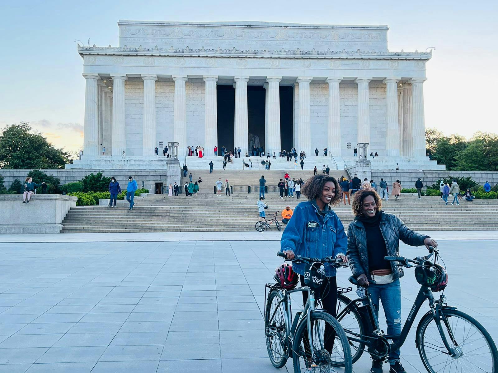 Two people standing with bicycles in front of the Lincoln Memorial, with other visitors on the steps.