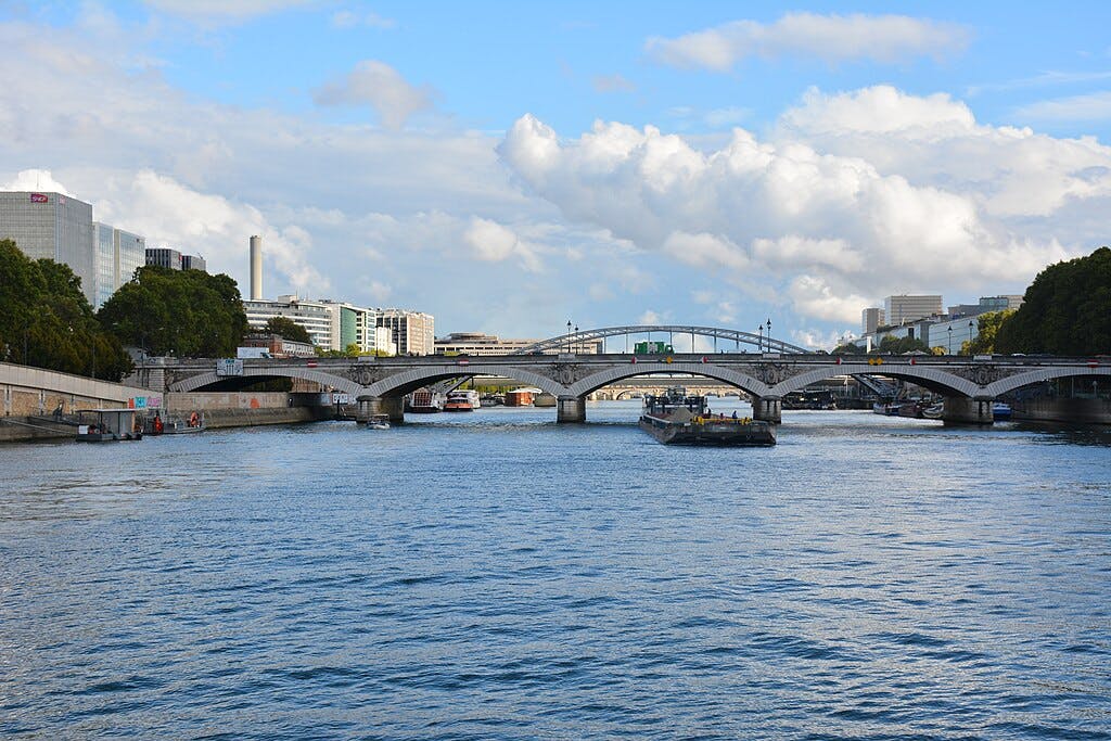 宽阔的河流在一系列华丽的桥梁下流淌。 在多云的蓝天下，两岸排列着建筑物和树木。