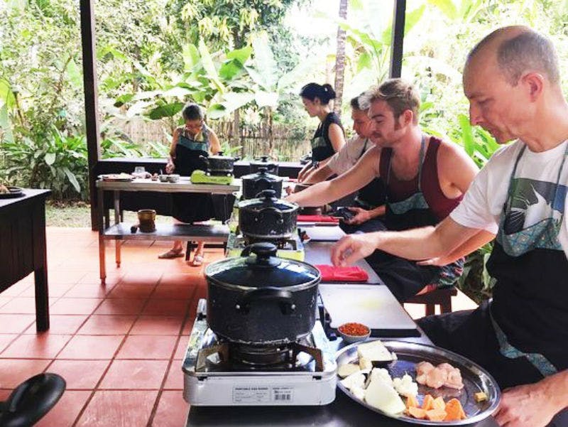 緑豊かな緑に囲まれた屋外ステーションで、エプロン姿の人たちが鍋やフライパンを使って料理をしています。