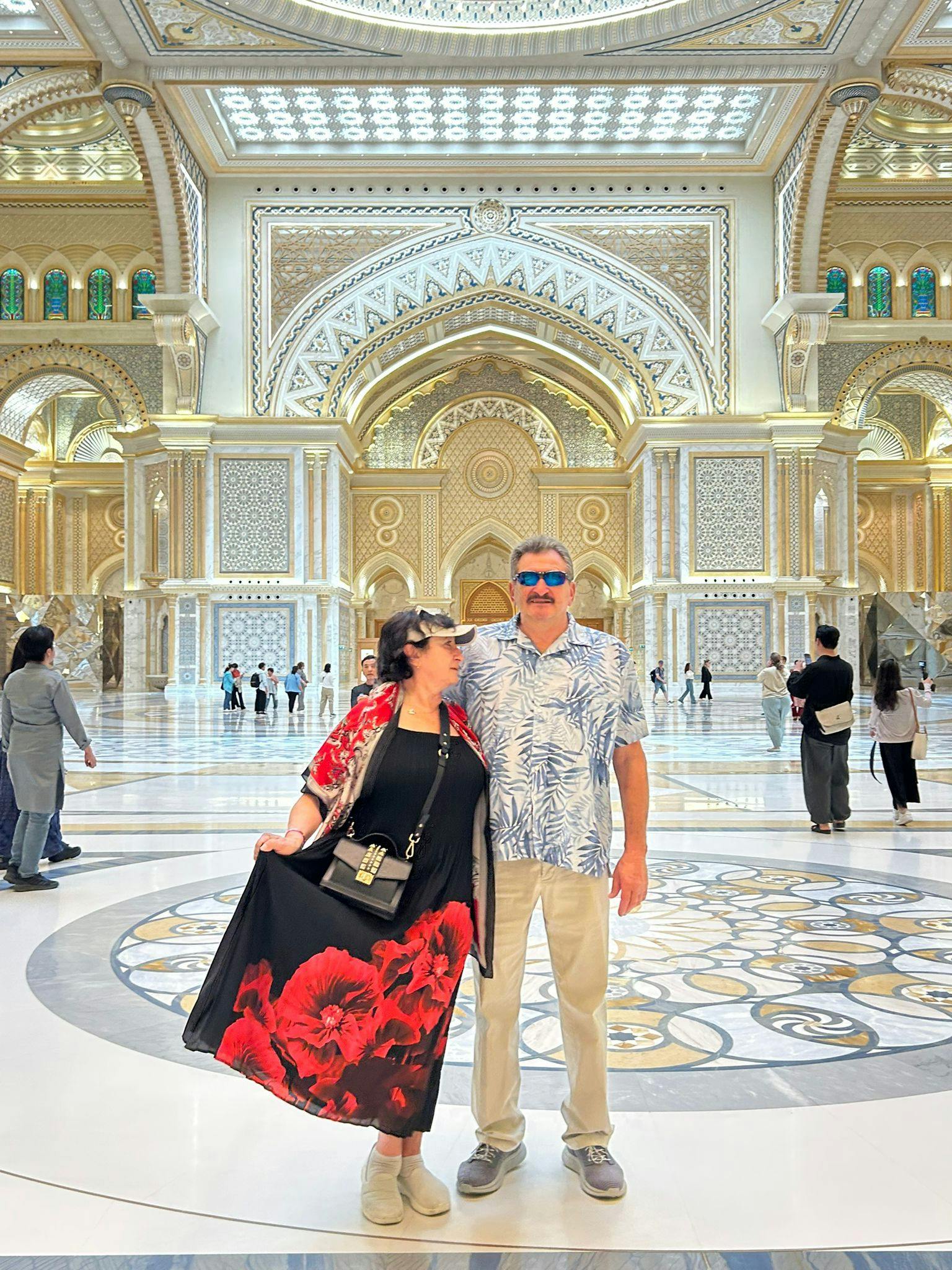 A man and woman pose in an ornate, gold and white interior with intricate patterns and arches, surrounded by other visitors.