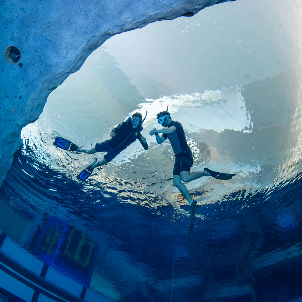 Δύο δύτες με στολές κατάδυσης και εξοπλισμό για κολύμβηση με αναπνευστήρα κολυμπούν υποβρύχια κοντά σε μια βραχώδη προεξοχή.