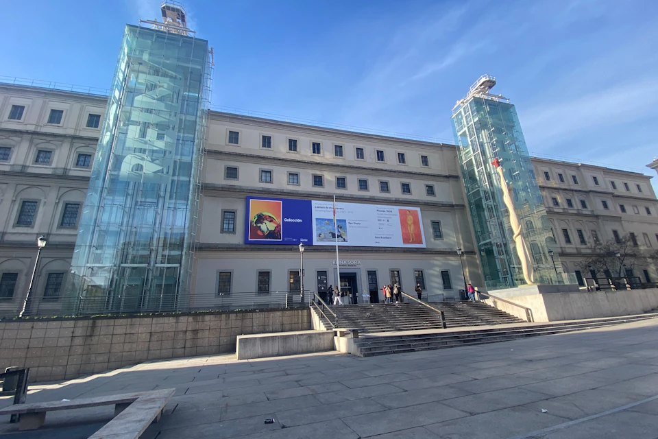 A rectangular building with banners, flanked by two glass elevator towers, and a staircase leading to its entrance.